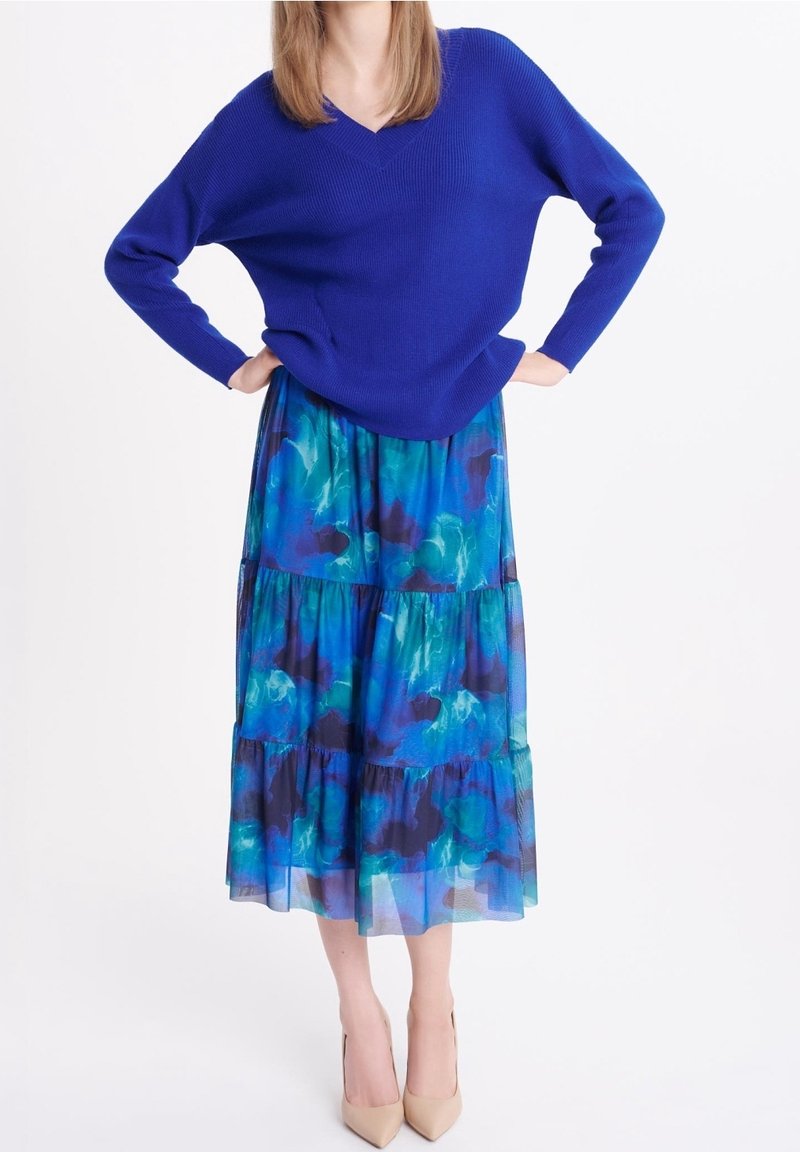 Femme portant un pull bleu vif à col en V et une jupe midi fluide à volants avec un motif bleu et vert, accompagnée de talons beiges.
