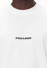 Bílý bavlněný tričko se černě tištěným logem "PEGADOR" uprostřed. Jednoduchý design s kulatým výstřihem, hladká textura.