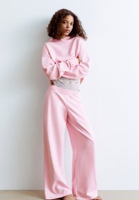 Felpa rosa con un fit rilassato e design cropped, abbinata a pantaloni della tuta rosa a gamba larga. Dettaglio in pizzo a strati visibile in vita.