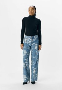 Un suéter de cuello alto negro, unos pantalones vaqueros de mezclilla azul claro con un patrón floral blanco, de corte amplio, combinados con unos zapatos de tacón negro.