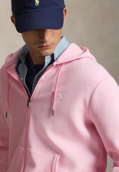 Mees kannab merev sinist nokamütsi, helesinist nööbiga särki ja helelillat lukuga kapuutsi, millel on rinnal väike tikitud logo, vaatab vasakule alla.