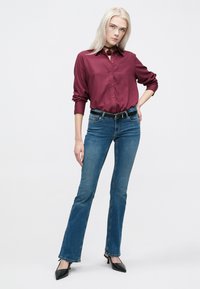 Camicia bordeaux con bottoni, jeans svasati blu, cintura nera e tacchi a punta neri. Tessuto morbido con colletto, vestibilità casual e stile denim classico.
