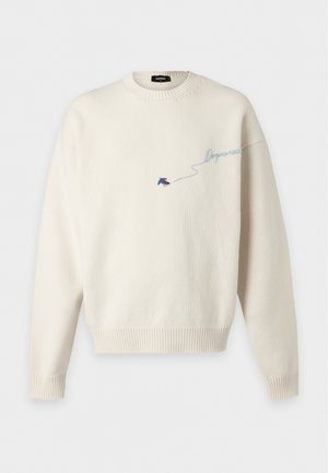 Jeune homme portant un pull crème avec une broderie bleue, un jean clair déchiré et des bottes foncées avec des lacets rouges, debout devant un fond blanc.