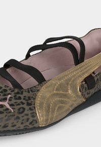 Κοντινή λήψη παπουτσιού Puma με λεοπάρ μοτίβο, χρυσές σουέντ λεπτομέρειες και μαύρα ελαστικά λουράκια στο πάνω μέρος.