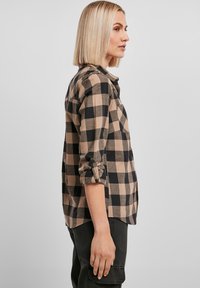 Chemise en flanelle à carreaux beiges et noirs avec manches retroussées, poche poitrine unique et texture douce, associée à un pantalon sombre.
