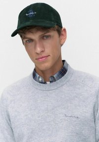 Grön kordfløjlskapa med broderad logotyp, bärs med en grå stickad tröja med rund hals över en blårutigt skjorta.