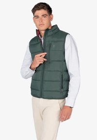Chaleco acolchado verde con cuello alto, cierre delantero y bolsillos laterales; la tela tiene una textura suave y presenta un forro interior burdeos.
