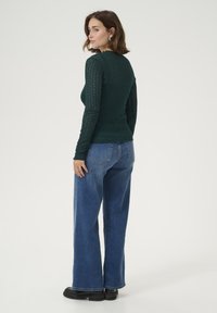 T-shirt à manches longues, tricoté en vert foncé avec un motif texturé, associé à un jean bleu à taille haute et jambes larges, et des chaussures à plateforme noires.