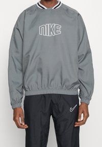 Pullover grigio Nike con cintura ed elastico ai polsi, caratterizzato da logo bianco sul petto e colletto a righe bianche e nere. Materiale morbido e liscio.