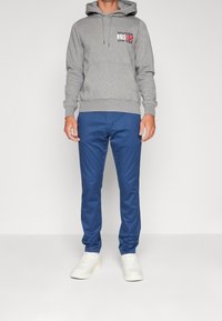 Felpa grigia con tasca frontale e cappuccio regolabile, caratterizzata dal logo "HILFIGER 1985 NEW YORK" in rosso e bianco. Abbinata a pantaloni blu e sneakers bianche.