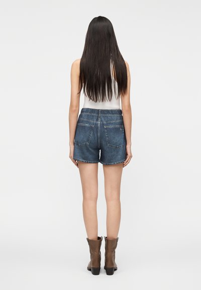 Shorts di denim a vita alta con orlo grezzo, in una tonalità di blu medio. Presentano tasche posteriori e una vestibilità comoda. Indossati con stivaletti marroni.