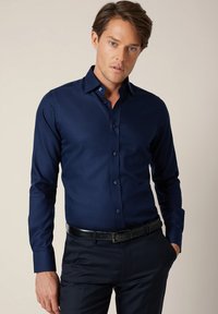 Granatowa koszula z długim rękawem wykonana z gładkiej tkaniny o dopasowanym kroju, z kontrastującymi guzikami i kołnierzem typu rozłożysty. Noszona z ciemnymi spodniami.