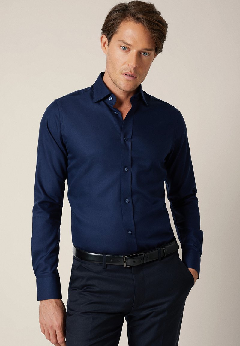 Granatowa koszula z długim rękawem wykonana z gładkiej tkaniny o dopasowanym kroju, z kontrastującymi guzikami i kołnierzem typu rozłożysty. Noszona z ciemnymi spodniami.