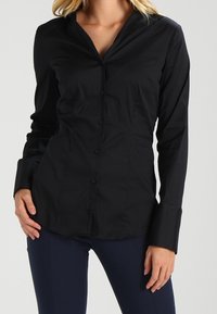 Chemise noire à manches longues avec un col pointu, dotée d'un design ajusté, d'une texture lisse et de boutons visibles sur le devant.