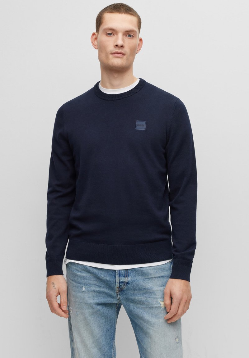 Marineblauwe gebreide trui met ronde hals, voorzien van een klein donker label op de borst en geribbelde boorden en onderrand. Gecombineerd met lichte spijkerbroeken.