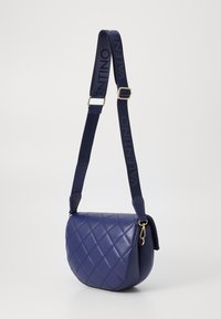Geantă crossbody din piele matlasată, de culoare bleumarin, cu accesorii aurii și o curea lată din material embosat cu "VALENTINO". Formă rotundă și textură moale.
