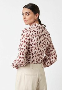 Frau mit dunklem Haar in einem tiefen Pferdeschwanz, die goldene Creolen, eine Langarmbluse mit Animal-Print und helle, bequeme Hochtaillen-Hosen trägt.
