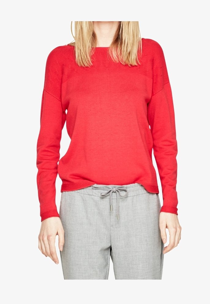 Pull en maille rouge à coupe décontractée, col côtelé et manches longues, associé à un pantalon gris clair à enfiler avec une taille élastiquée et un cordon de serrage.