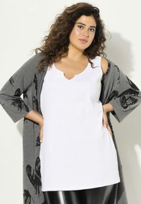 Witte mouwloze top met een V-hals, gecombineerd met een grijze cardigan met zwart vlinderprint. Model draagt zwarte legging.
