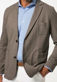 Grauer maßgeschneiderter Blazer mit einer Brusttasche, weicher Textur und Zwei-Knopf-Verschluss vorne. Kombiniert mit einem hellblauen, karierten Hemd darunter.