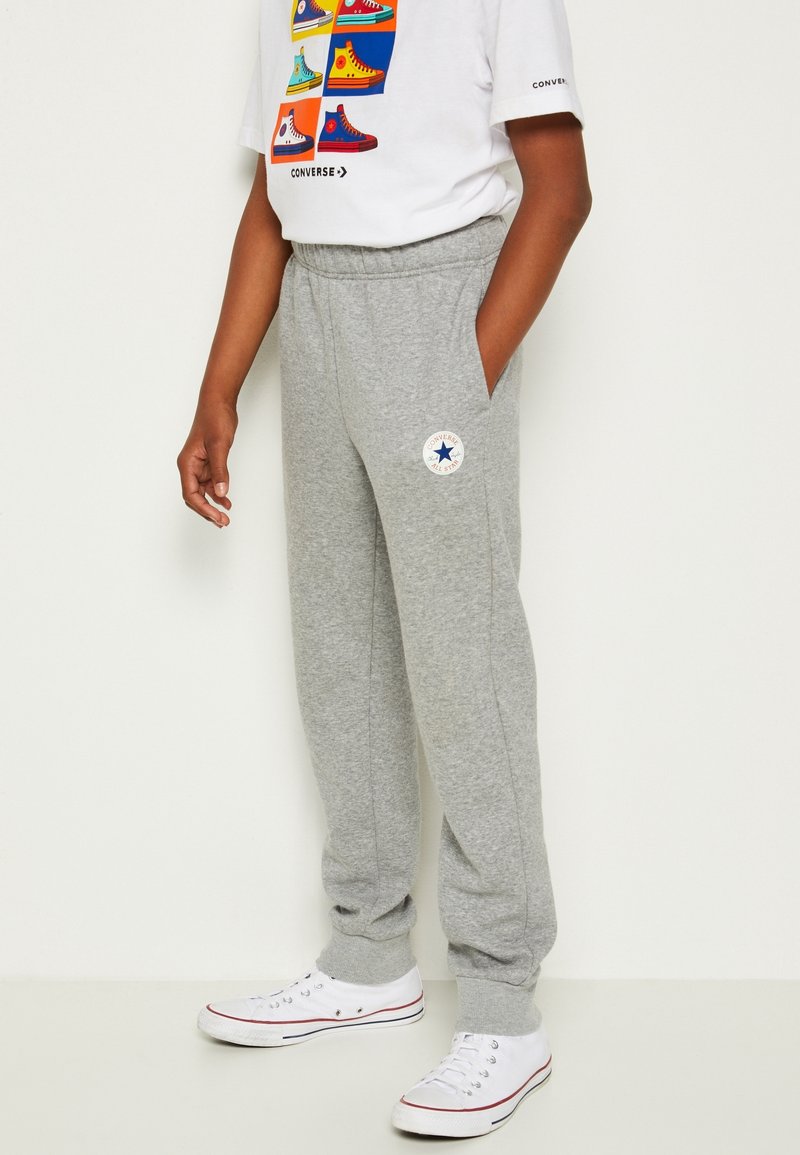 Grå sweatpants med elastisk talje, sidelommer, ribbede hæmper og et Converse-logo-patch på venstre lår. Båret med hvide sneakers.