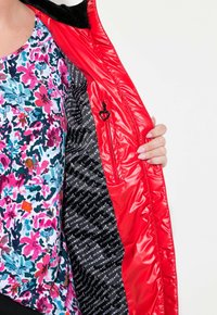 Chaquetón rojo acolchado con forro negro que presenta un patrón de logotipos; exterior suave y brillante. Una parte de una blusa floral colorida es visible.
