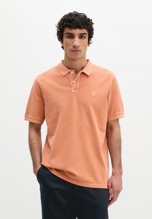Jonge man met krullend haar, gekleed in een zalmroze poloshirt met korte mouwen en een donkere broek, staand tegen een effen witte achtergrond.