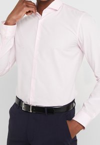 Camicia a maniche lunghe rosa pallido con colletto a bottoni, texture liscia e bottoni bianchi. Abbinata a pantaloni scuri e una cintura nera.