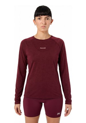 Žena s krátkými černými vlasy, oblečená v přiléhavém bordovém tričku s dlouhým rukávem a odpovídajících šortkách, stojící na bílém pozadí.