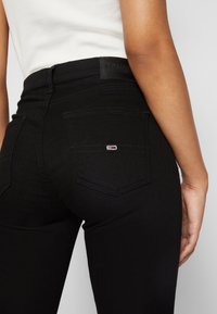 Jeans negros de tiro alto con un diseño ajustado. Cuenta con un parche de cuero con logo, dos bolsillos traseros y detalles de costura en contraste.