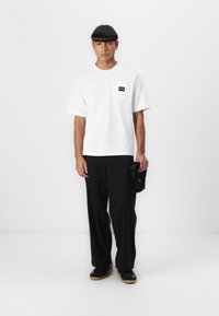 White short-sleeve T-shirt with a chest pocket and black pants. Model holds a small black bag. Shoes are black with a gum sole.