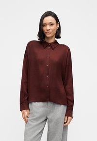 Burgunderfarvet langærmet skjorte med krave, knappet front og afslappet pasform. Glat tekstur og let oversized design, kombineret med grå bukser.