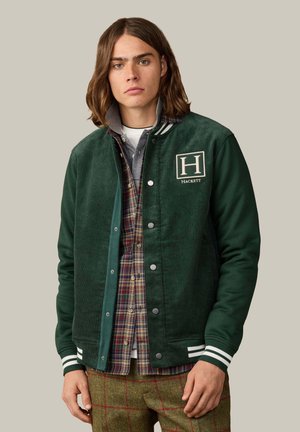 Junger Mann mit schulterlangen Haaren trägt eine grüne Collegejacke über einem karierten Hemd und grünen karierten Hosen und steht vor einem einfarbigen Hintergrund.