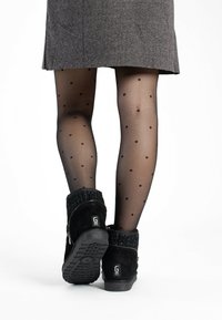 Botines de ante negros con un puño de punto texturizado, usados con medias negras de lunares transparentes, sobre un fondo claro.