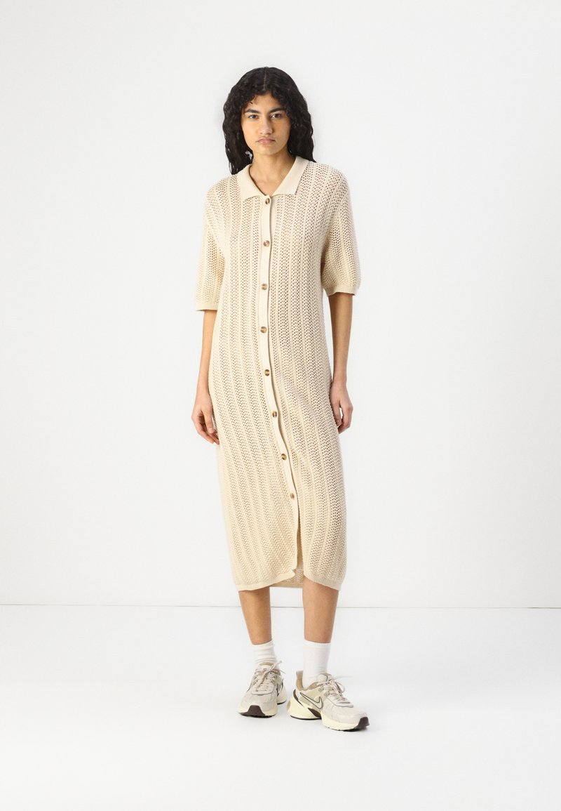 Gehaakte beige jurk met korte mouwen, knoopsluiting aan de voorkant en een geribbelde structuur, tot halverwege de kuit. Gecombineerd met beige sneakers.
