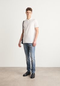Camiseta polo blanca de algodón con mangas cortas, combinada con jeans de color azul claro. Zapatos negros con detalles texturados. El modelo sostiene un bolso de hombro.