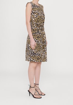 Femme portant une robe sans manches longueur genou à imprimé léopard avec taille froncée, associée à des sandales noires à talons hauts et brides, debout de profil.