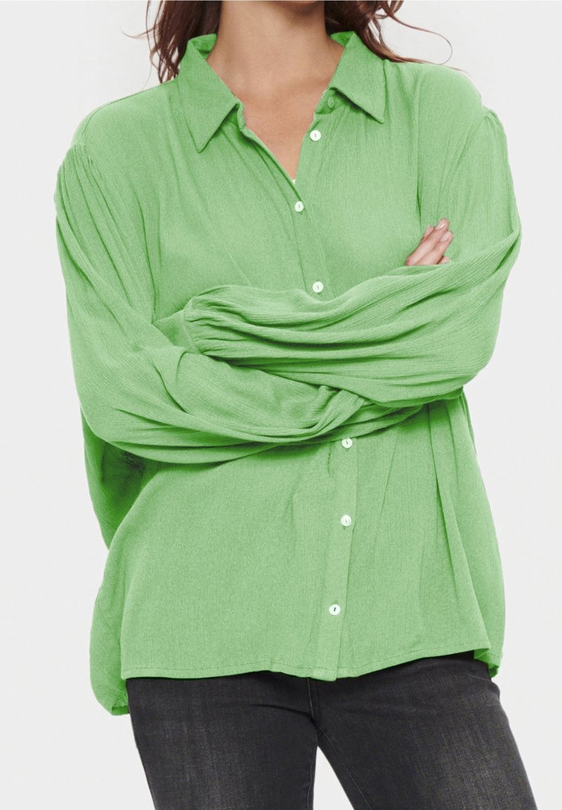 Blouse verte clair à manches longues avec un devant à boutons, un col et une coupe ample. Tissu texturé avec des plis subtils sur les manches.