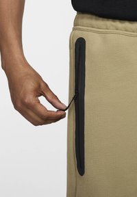 Kaki joggers med en svart dragkedjeficka. Hand som drar i snörjusteraren, vilket visar på ett mjukt material och en elegant design.