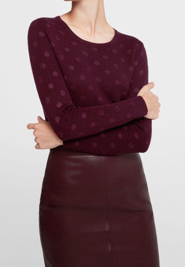 Personne portant un pull à manches longues bordeaux avec des points texturés et une jupe en cuir bordeaux assortie, bras croisés.