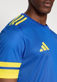 Maglietta sportiva blu realizzata in materiale liscio. Presenta strisce gialle sulle maniche e un design con tre strisce sulla spalla. Scollo rotondo.