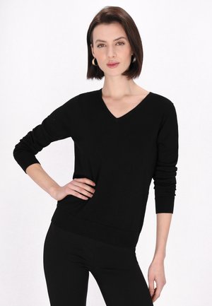 Kvinde med kort brunt hår iført guldbøjleøreringe, en sort V-hals sweater og sorte bukser, poserer med den ene hånd på hoften.
