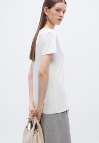 Λευκός, κοντομάνικος T-shirt από βαμβακερό ύφασμα, συνδυασμένο με γκρι φούστα. Ένα άτομο κρατά μια ανοιχτό μπεζ τσάντα με φερμουάρ.