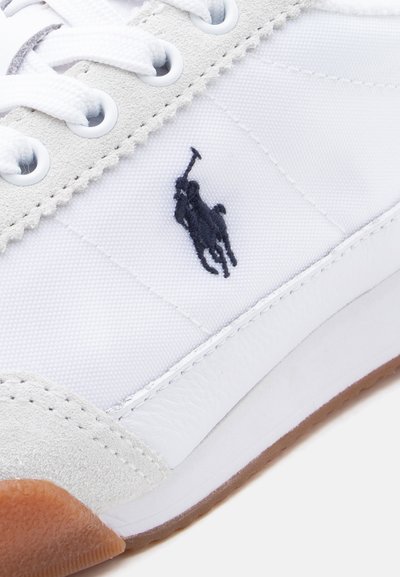 Hvit joggesko med semskede detaljer, flate lisser, gummisåle og en brodert marineblå logo. Har teksturert stoff og glatte skinn-detaljer.