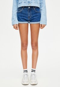 Persona de pie que lleva pantalones cortos de mezclilla azul de dos tonos, chaqueta de mezclilla azul claro, zapatillas blancas y calcetines blancos con rayas negras.