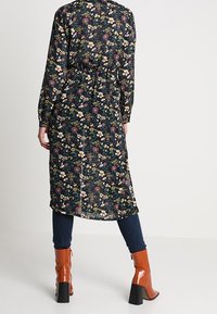 Lång svart blommig klänning med elastisk midja och knapppdetaljer, med färgglada blommor och en flödande silhuett, matchad med orange klackstövlar.