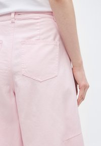 Γυναίκα που φοράει ροζ τζιν σορτς με πίσω τσέπες, στέκεται μπροστά σε απλό λευκό φόντο, με το δεξί χέρι χαλαρό στο πλάι.