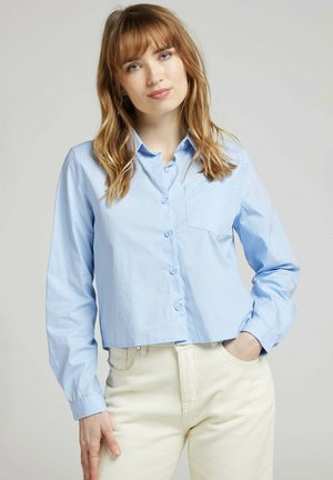 Chemise courte bleu clair à boutons, à manches longues et avec une poche poitrine présentant un détail à bord festonné. Tissu lisse, coupe décontractée.
