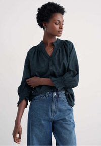 Žena s krátkymi kučeravými vlasmi m�á na sebe tmavozelenú kostkovanú blúzku s volánmi na rukávoch a vysokými modrými džínsami, stojí pred jednoduchým pozadím.