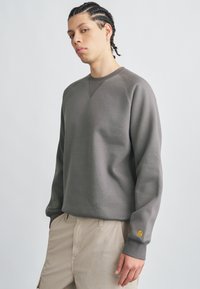 Gray sweatshirt with a ribbed texture, crew neckline, and raglan sleeves. Features a small yellow logo on the cuff and a relaxed fit.